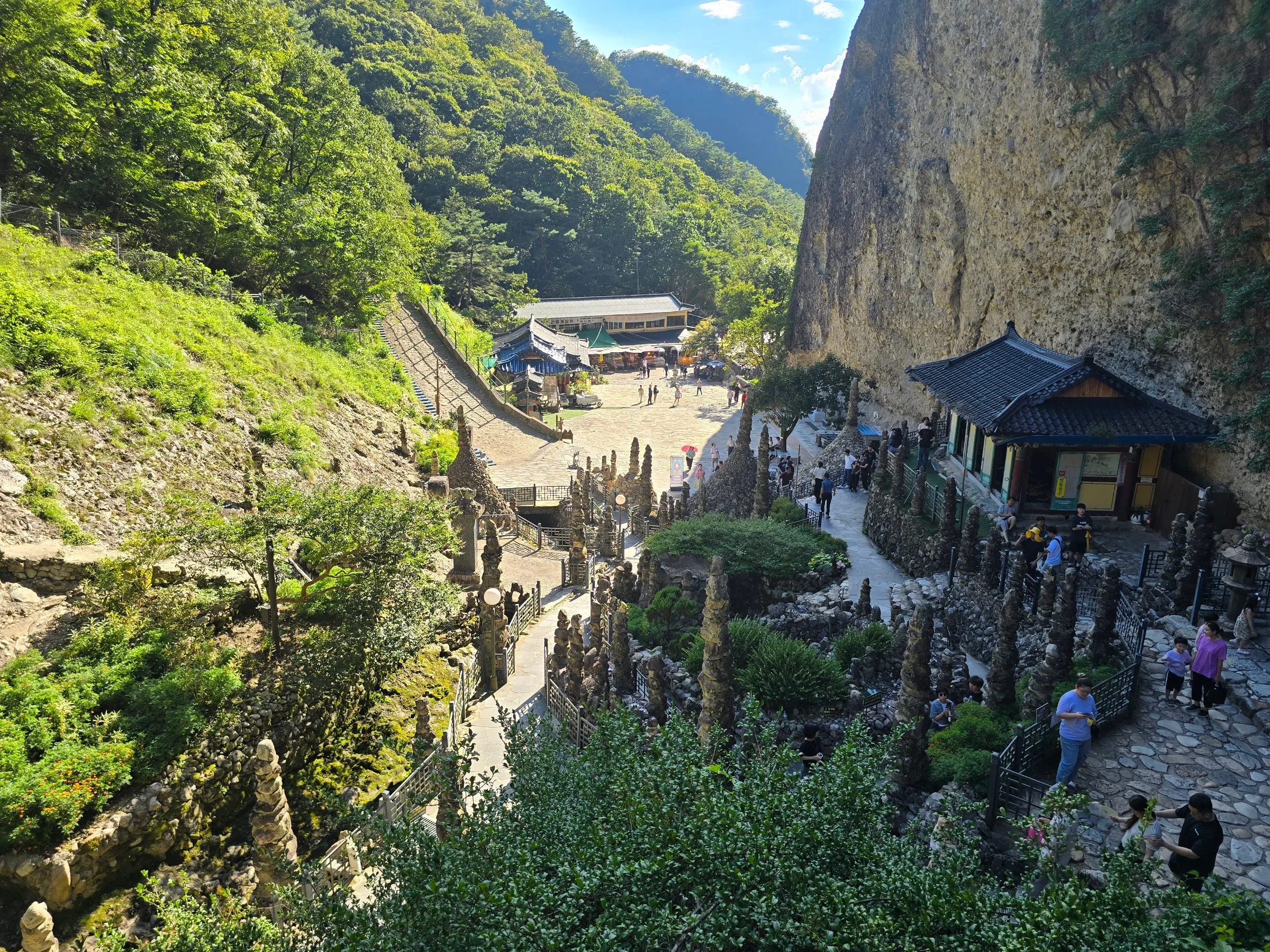Visitors walk among rows of hand-stacked stone pagodas below a tall rocky cliff, with a tile-roofed hall and forested green mountains behind.