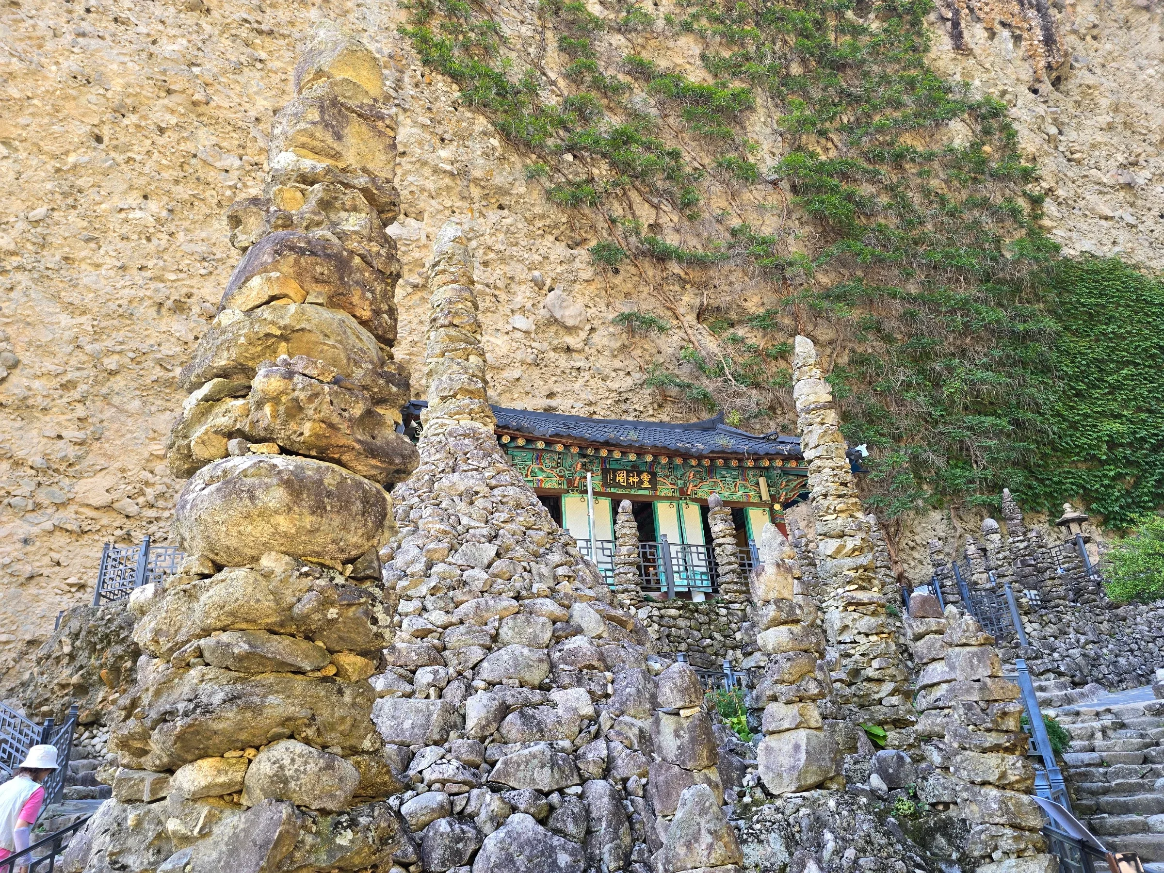 Hand-stacked stone pagodas stand before a tiled-roof hall with a 靈神閣 sign, beneath a steep conglomerate cliff with climbing ivy.