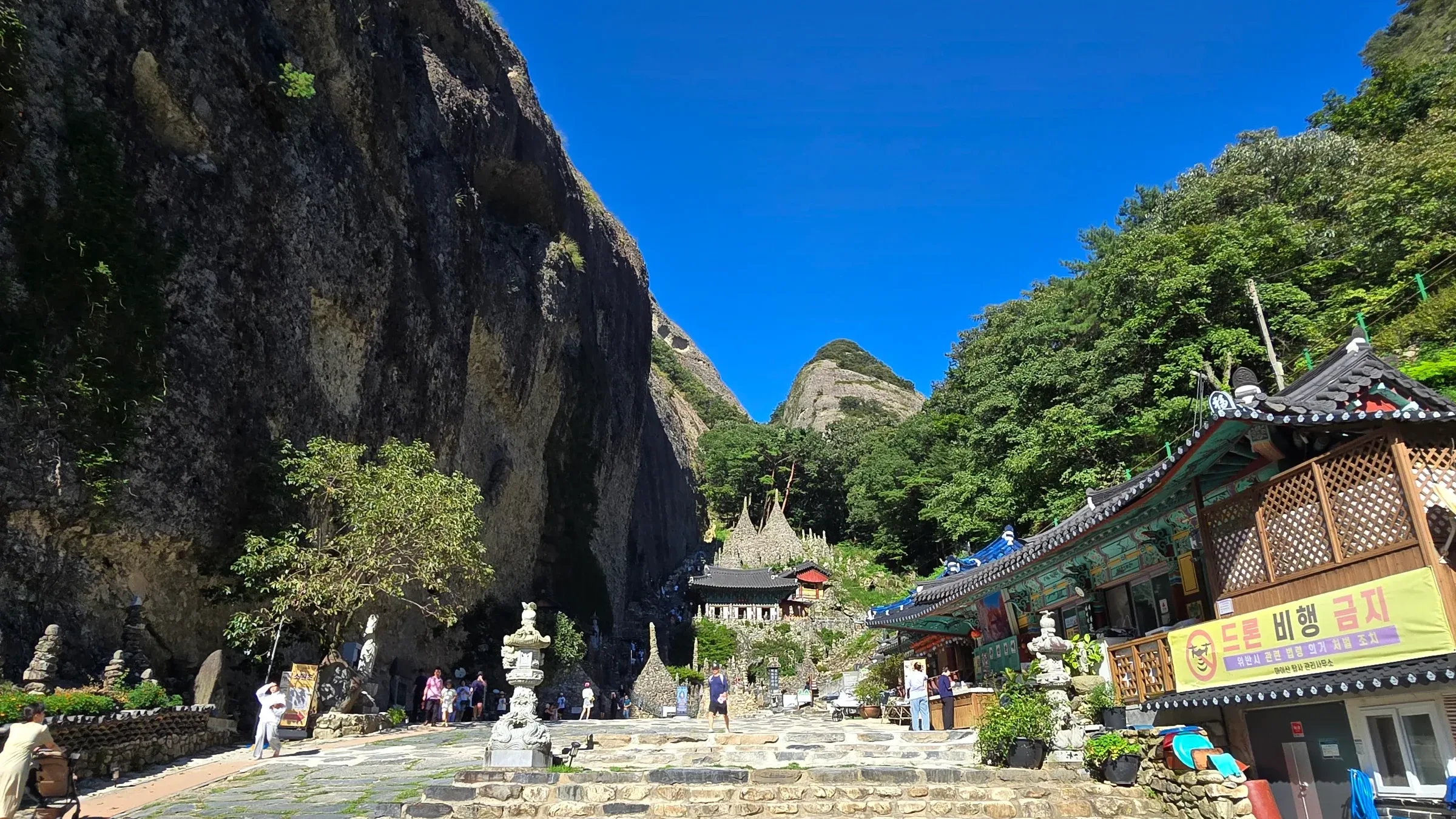 Stone-paved courtyard with stone lanterns and steps beneath a huge rock cliff, with stacked stone towers and a temple hall ahead and a banner reading 드론 비행 금지.