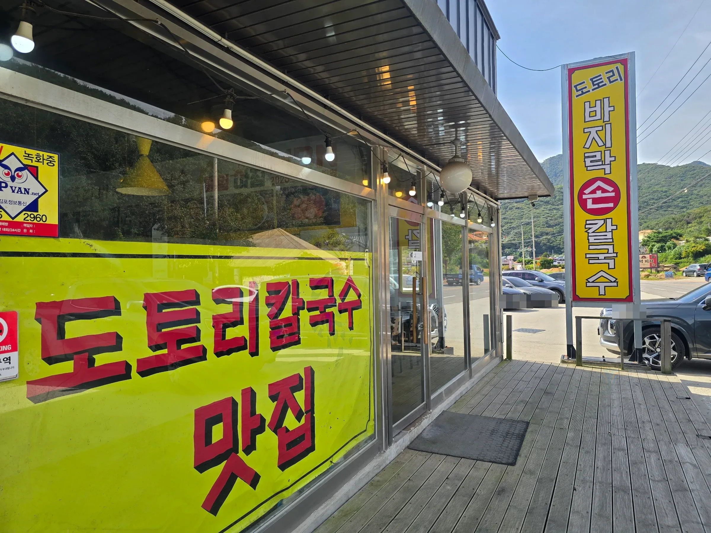 A glass-fronted restaurant with bright yellow Korean signs, a wooden deck, parked cars, and green hills beyond.