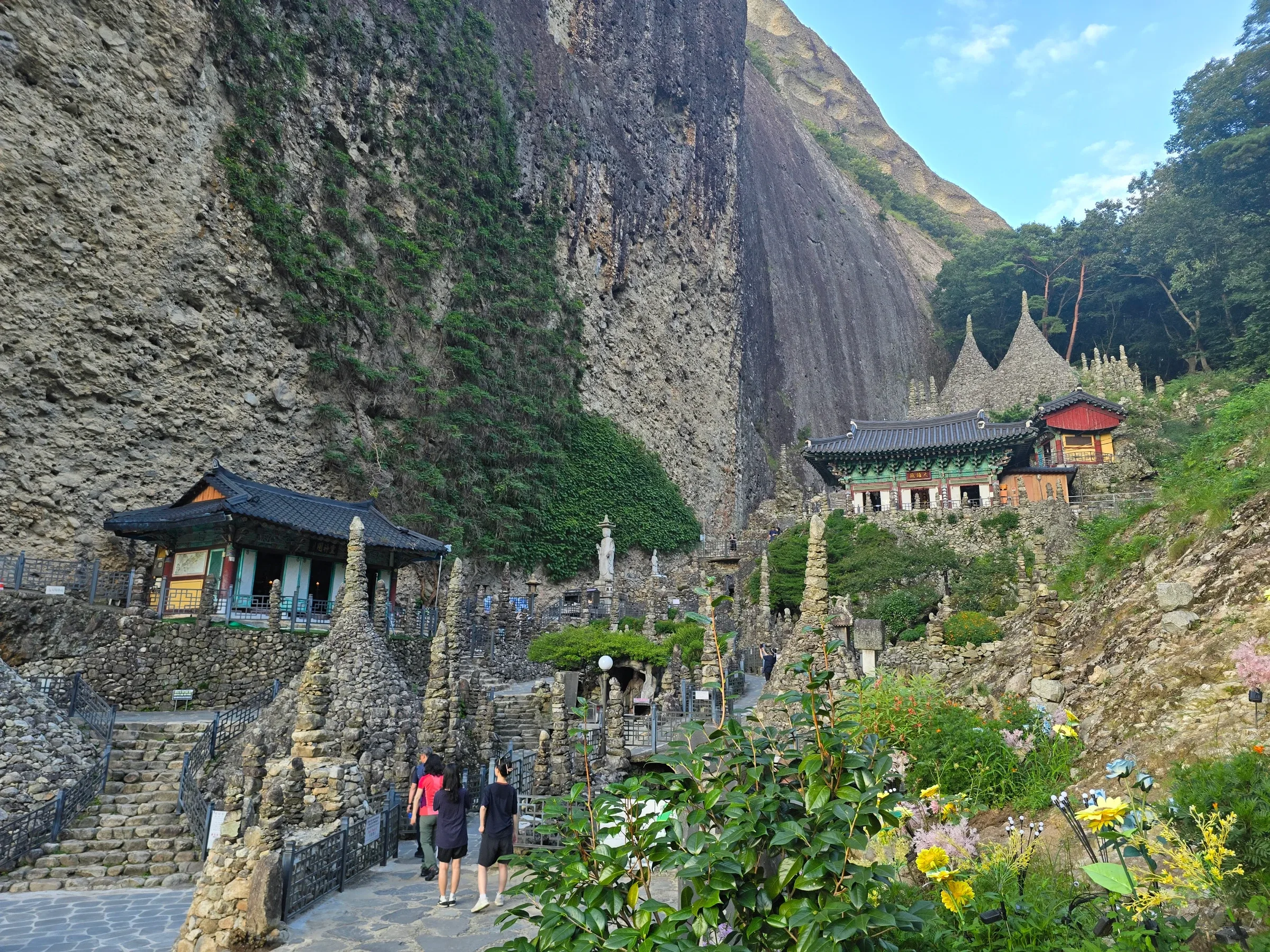 Visitors walk a stone path among hand-stacked stone pagodas toward a tiled-roof temple hall beneath a huge sheer rock cliff.