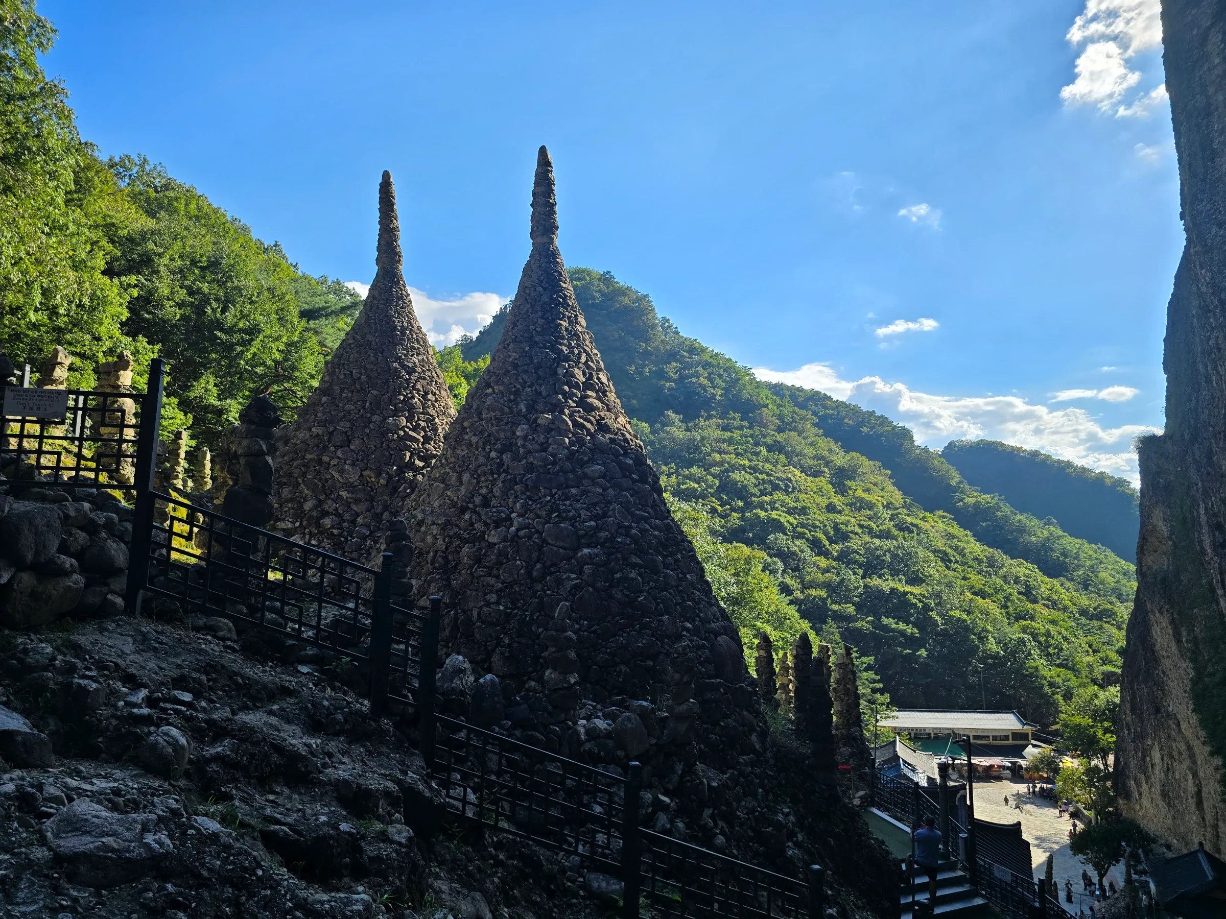Two tall conical stone pagodas of hand-stacked rocks rise beside a black railing, with forested green mountains, a blue sky and a temple courtyard below.