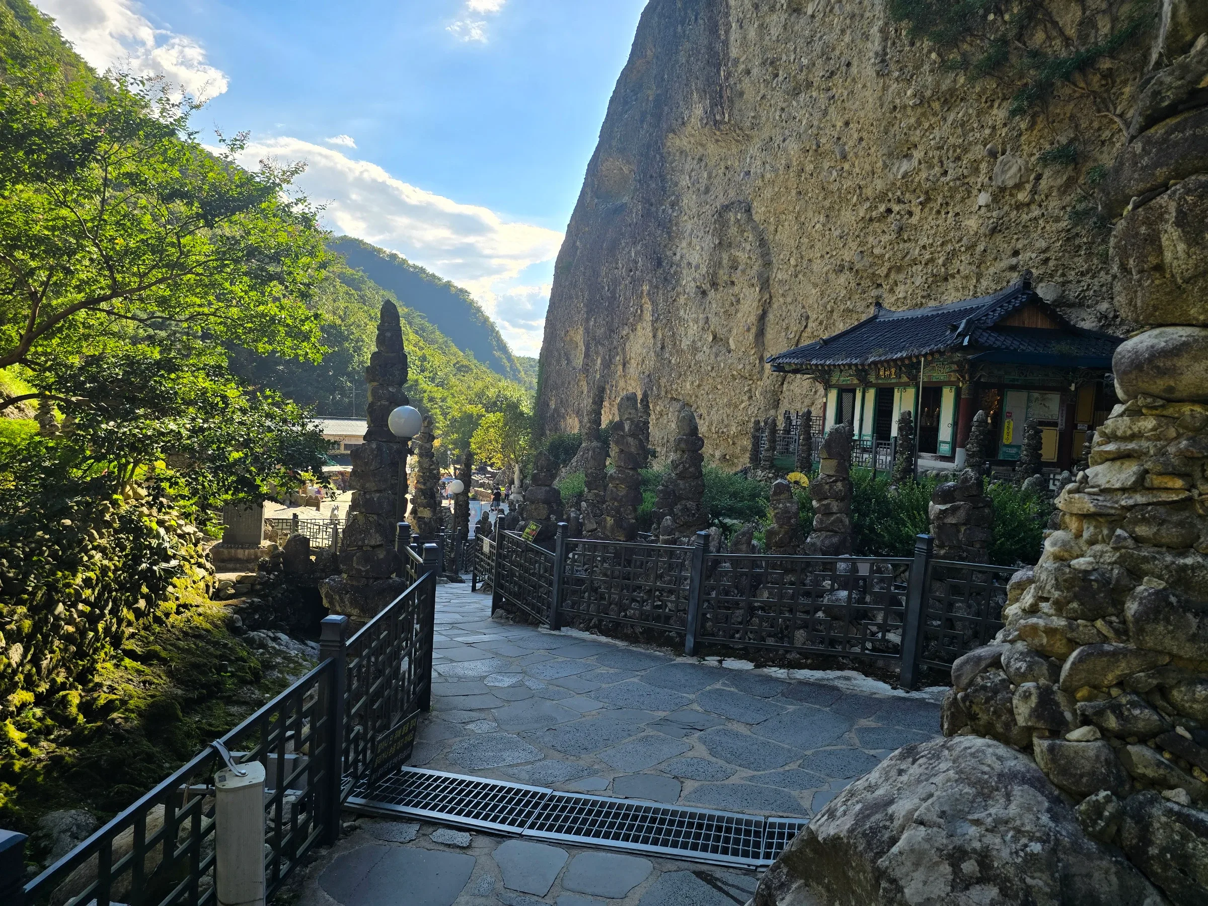 Stone-paved path with black railings winds past hand-stacked stone pagodas and a tile-roofed hall beneath a huge rock cliff in a green valley.