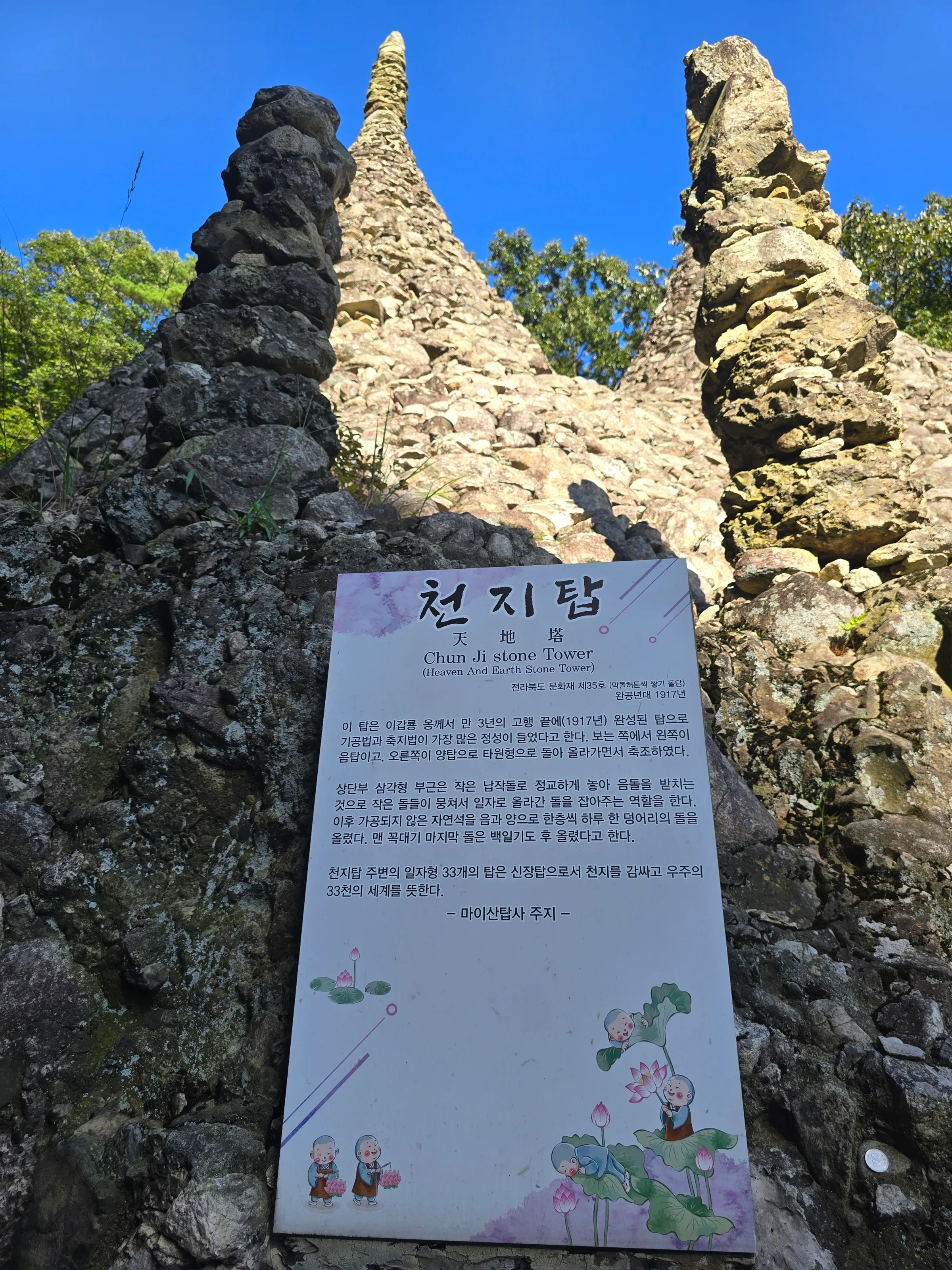 Two tall hand-stacked stone pagodas rise toward a blue sky above a white Korean sign titled Chun Ji stone Tower , set on a rock face.