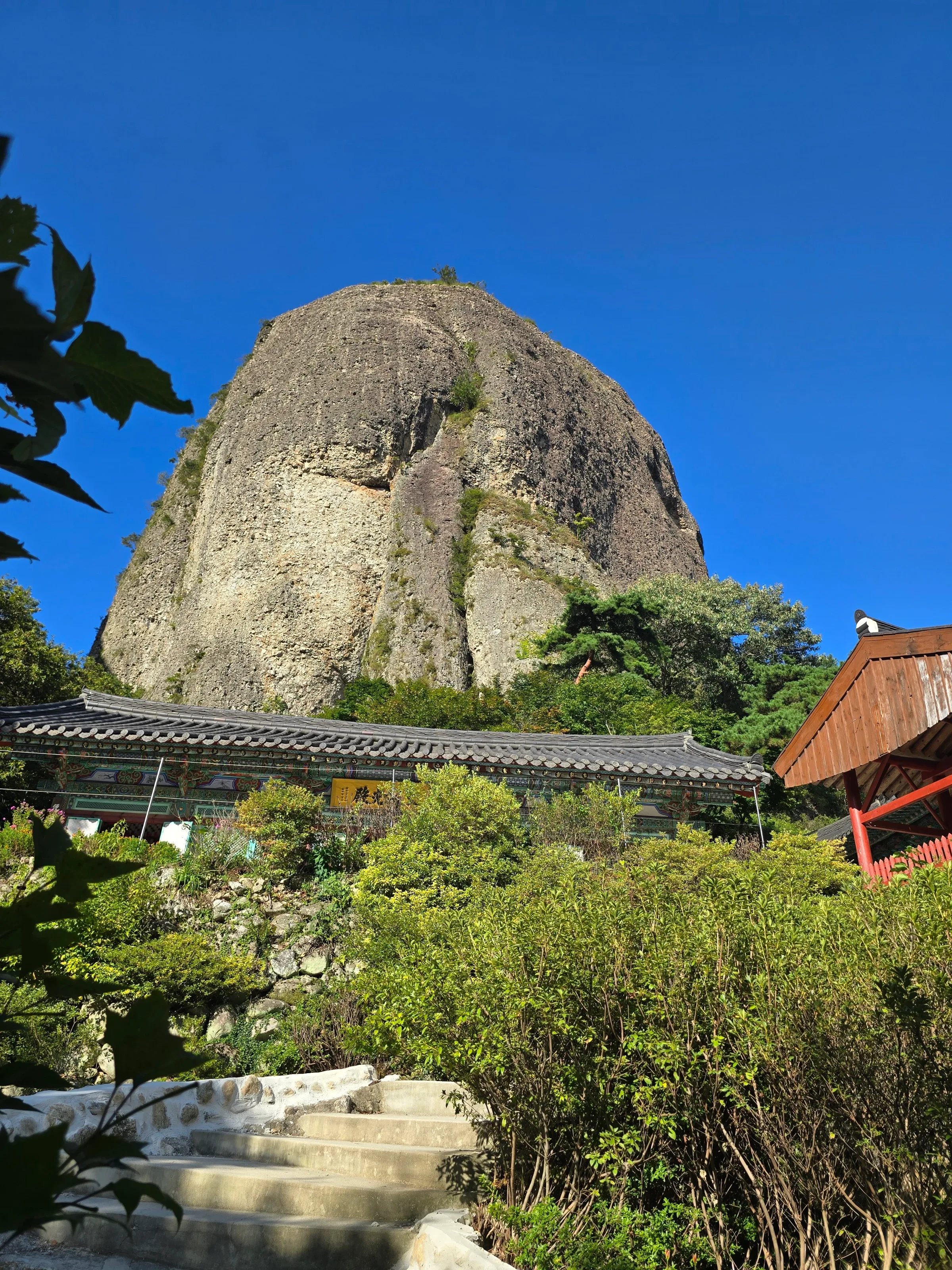 Looking up from stone steps and green shrubs at a traditional tile-roofed hall beneath a huge rounded rock peak against a deep blue sky.
