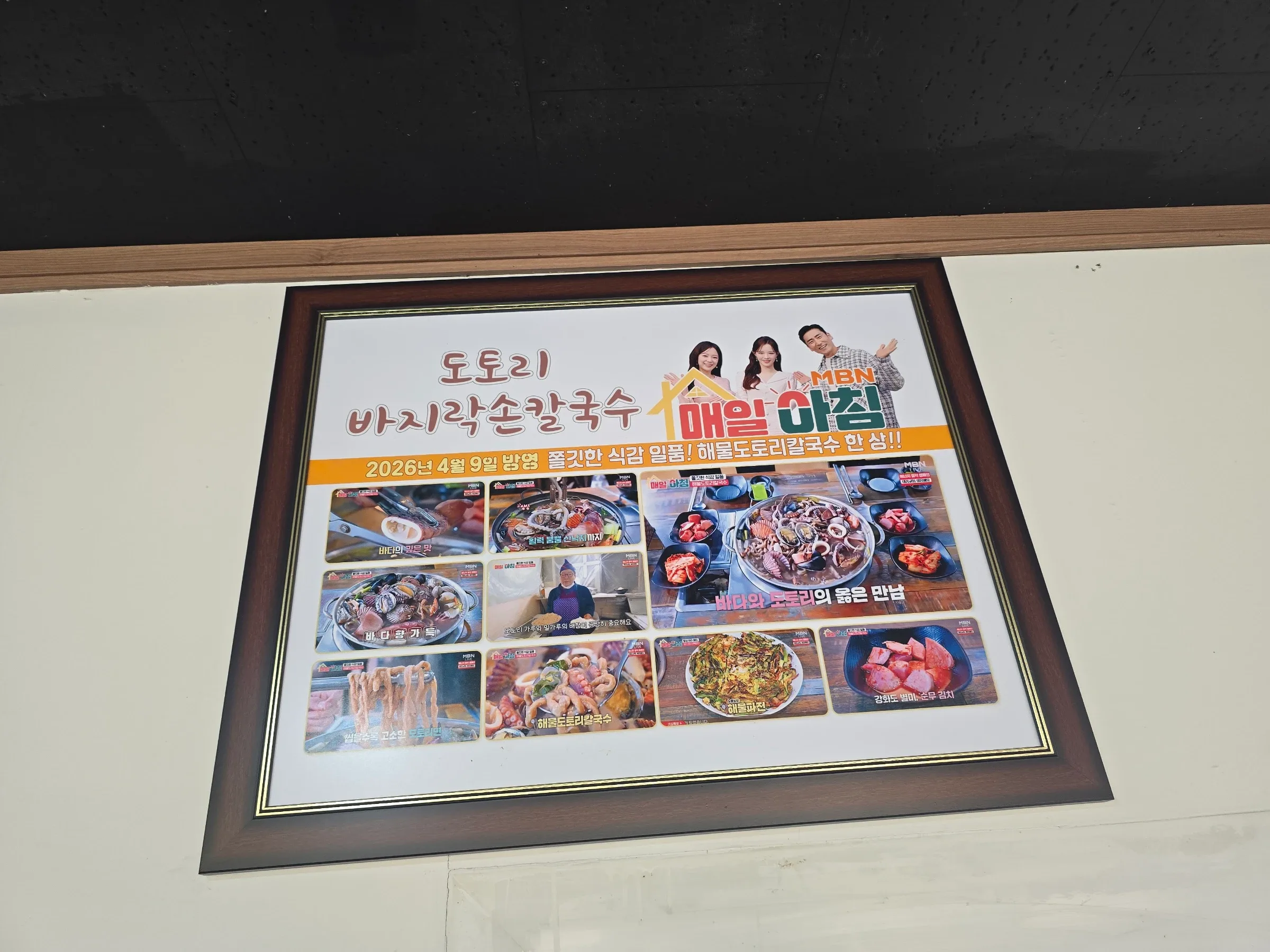 A framed Korean poster shows seafood noodle dishes, side dishes, and three TV presenters.