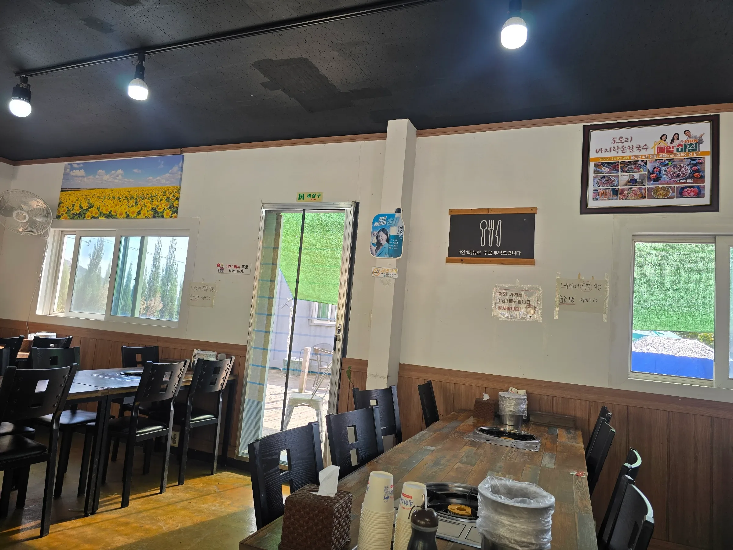 A restaurant dining room with wooden tables, black chairs, ceiling lights, windows, and framed food posters.