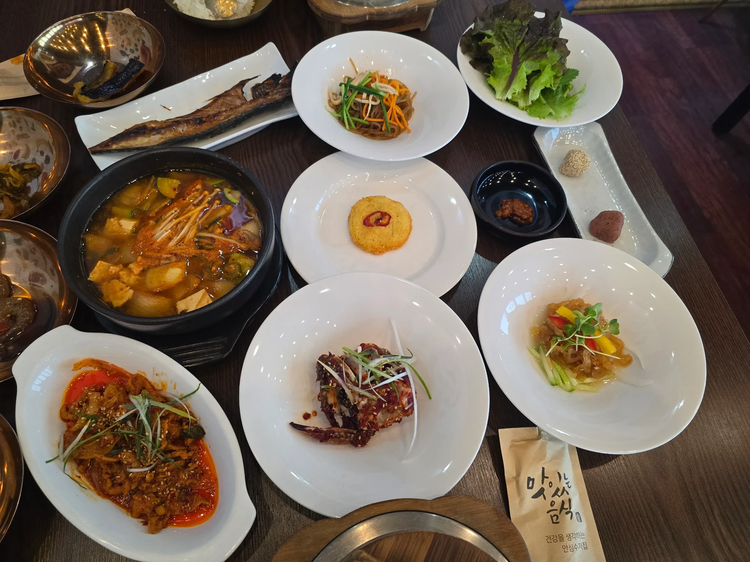 Korean hanjeongsik table with a bubbling tofu stew, grilled fish, japchae, spicy pork, seasoned crab, jellyfish salad, lettuce and brass side bowls