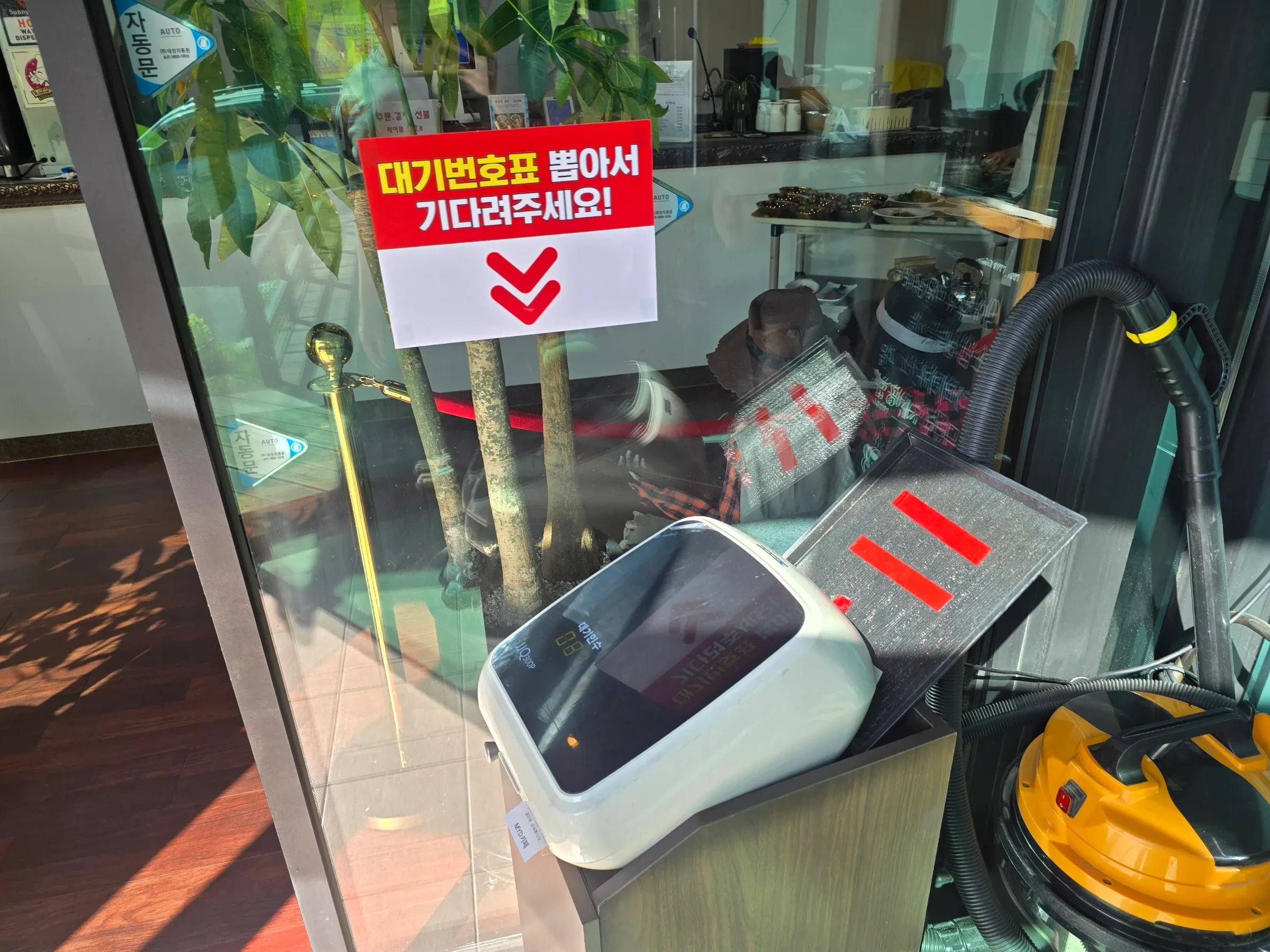 Behind a glass door, a red sign in Korean tells customers to take a waiting number, above a white ticket machine with a yellow vacuum cleaner beside it.