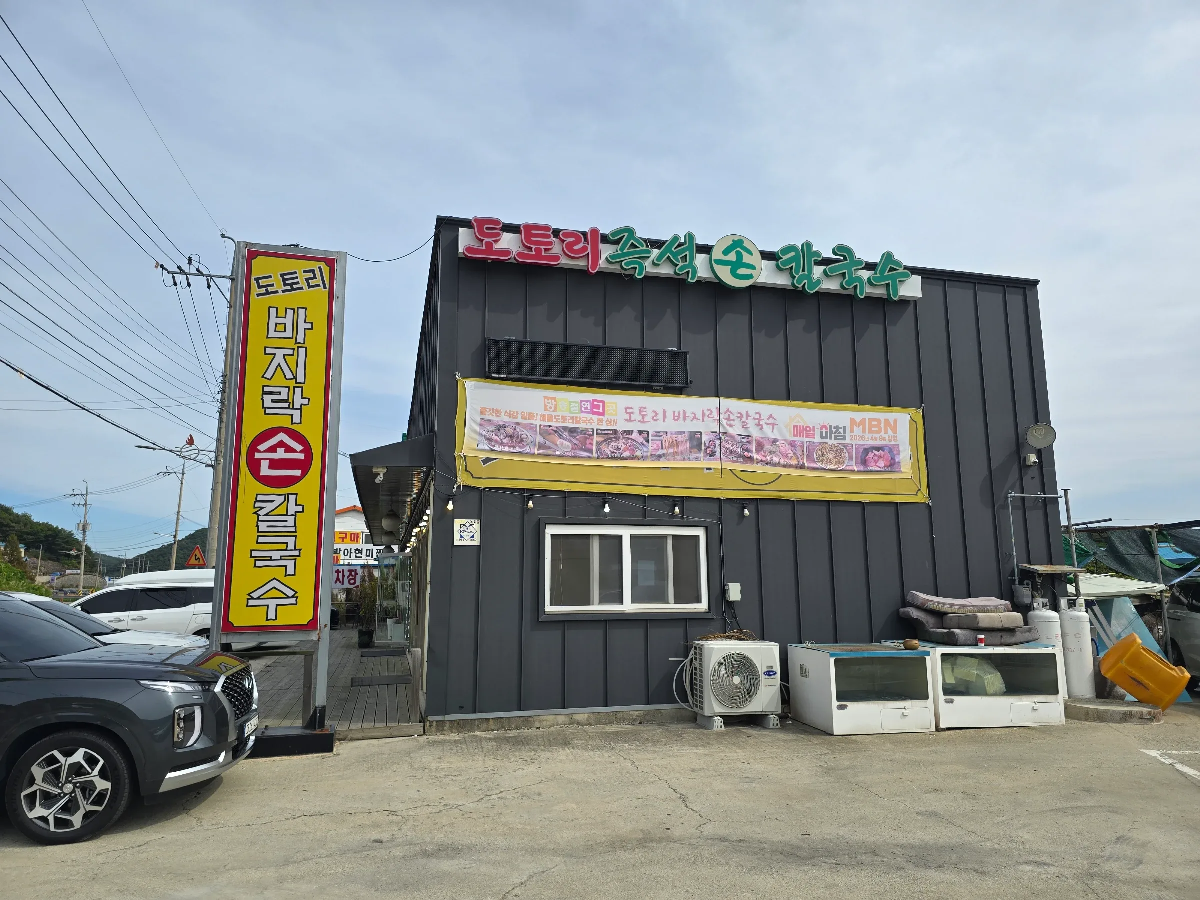 A dark-sided restaurant with colorful Korean signs and a yellow food banner stands beside parked cars.