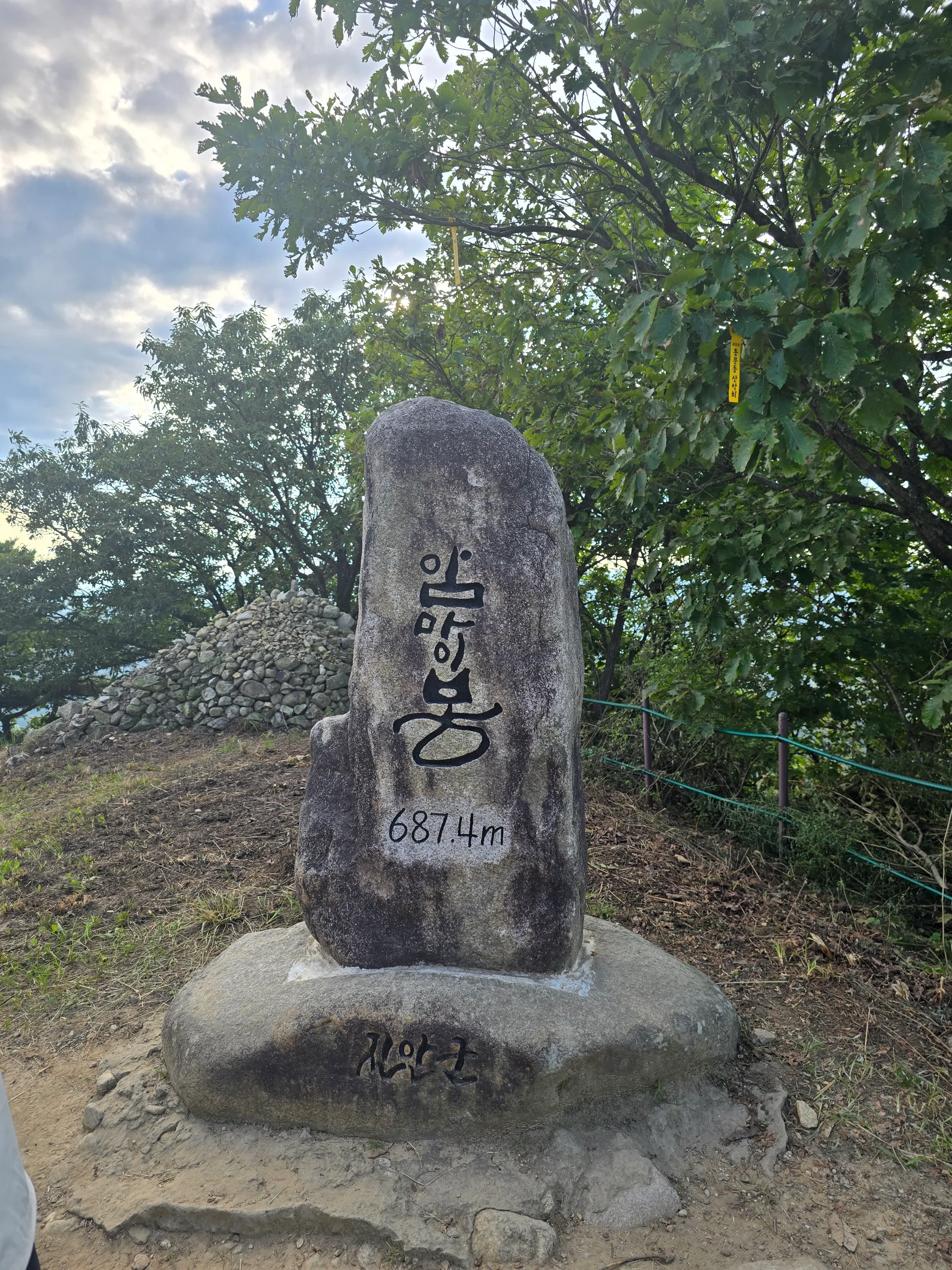 Tall carved granite summit marker reading 687.4m on a rock base, with a stone cairn, green trees, a green rope fence and cloudy sky behind.