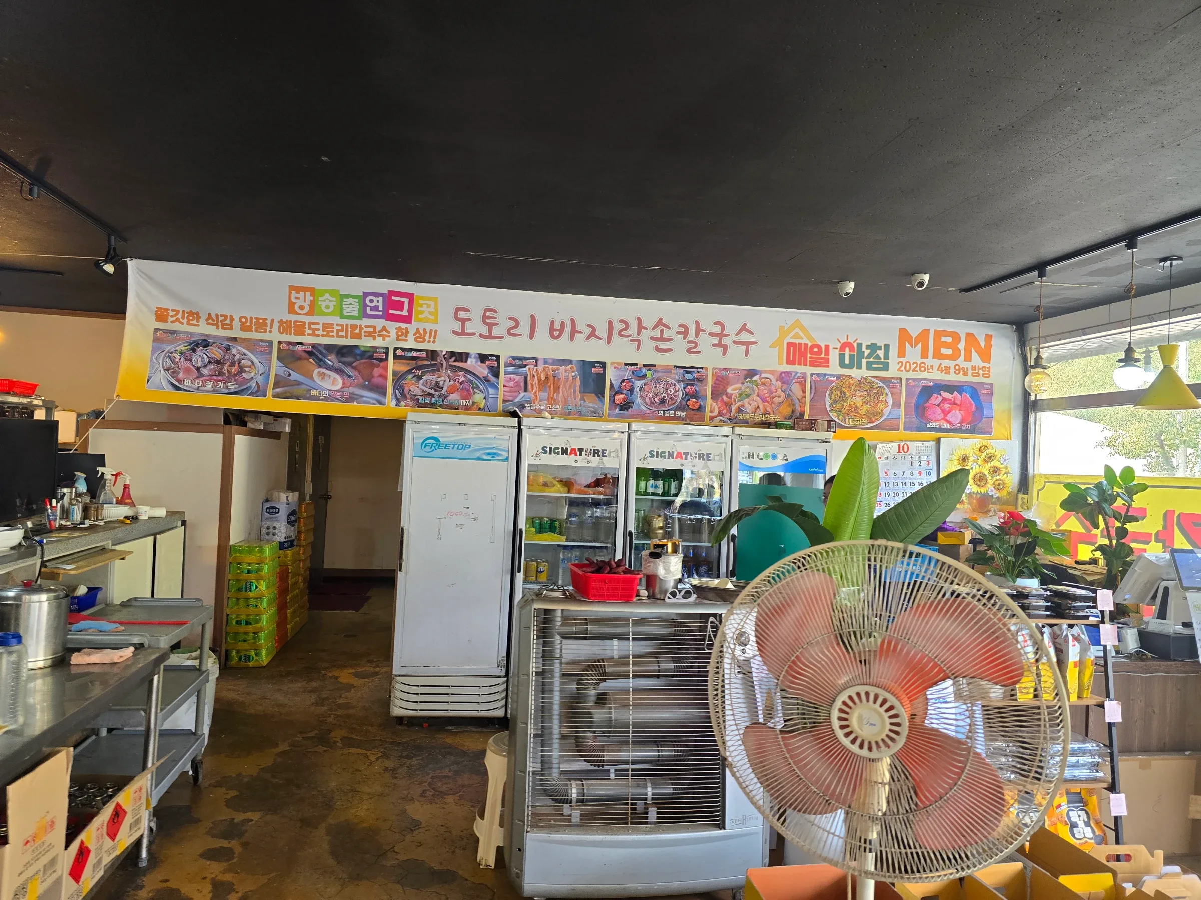 A restaurant interior with refrigerators, a large fan, potted plants, and a Korean banner featuring seafood noodle dishes.
