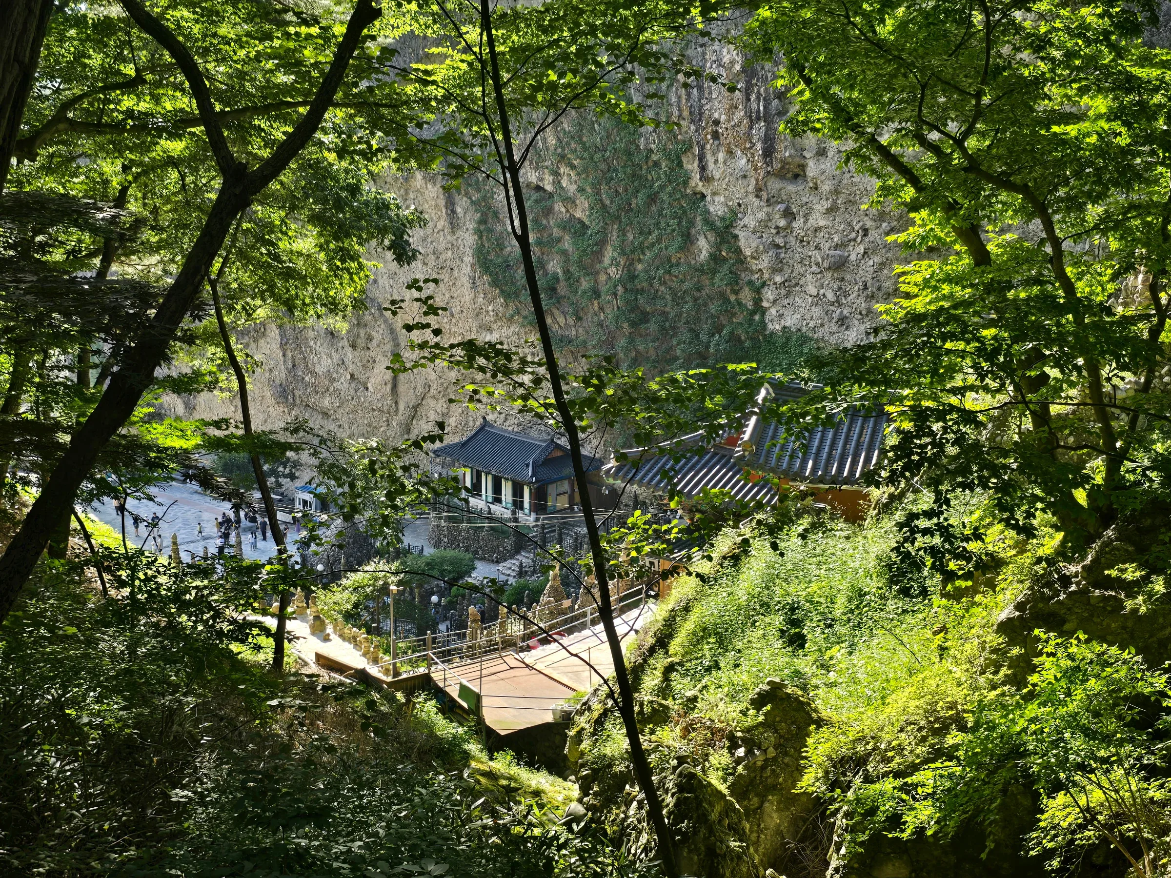 Green foliage frames a view down to a tiled-roof temple hall and a plaza with visitors, below a steep rock cliff, with small stacked stone towers lining the path.