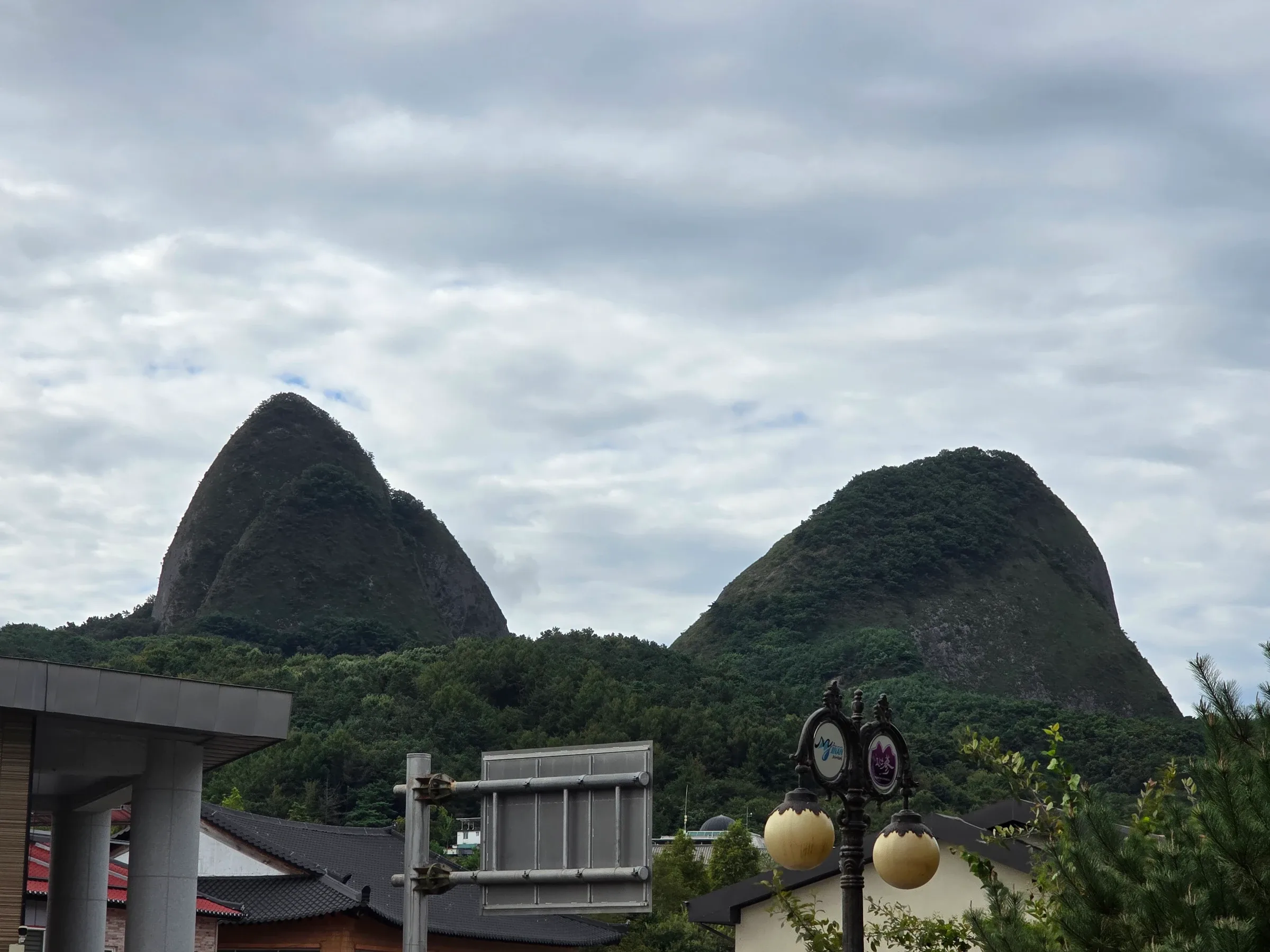Two steep, rounded green mountain peaks rise under a cloudy sky behind a lamp post with globe lights, a blank signboard and tiled-roof buildings.