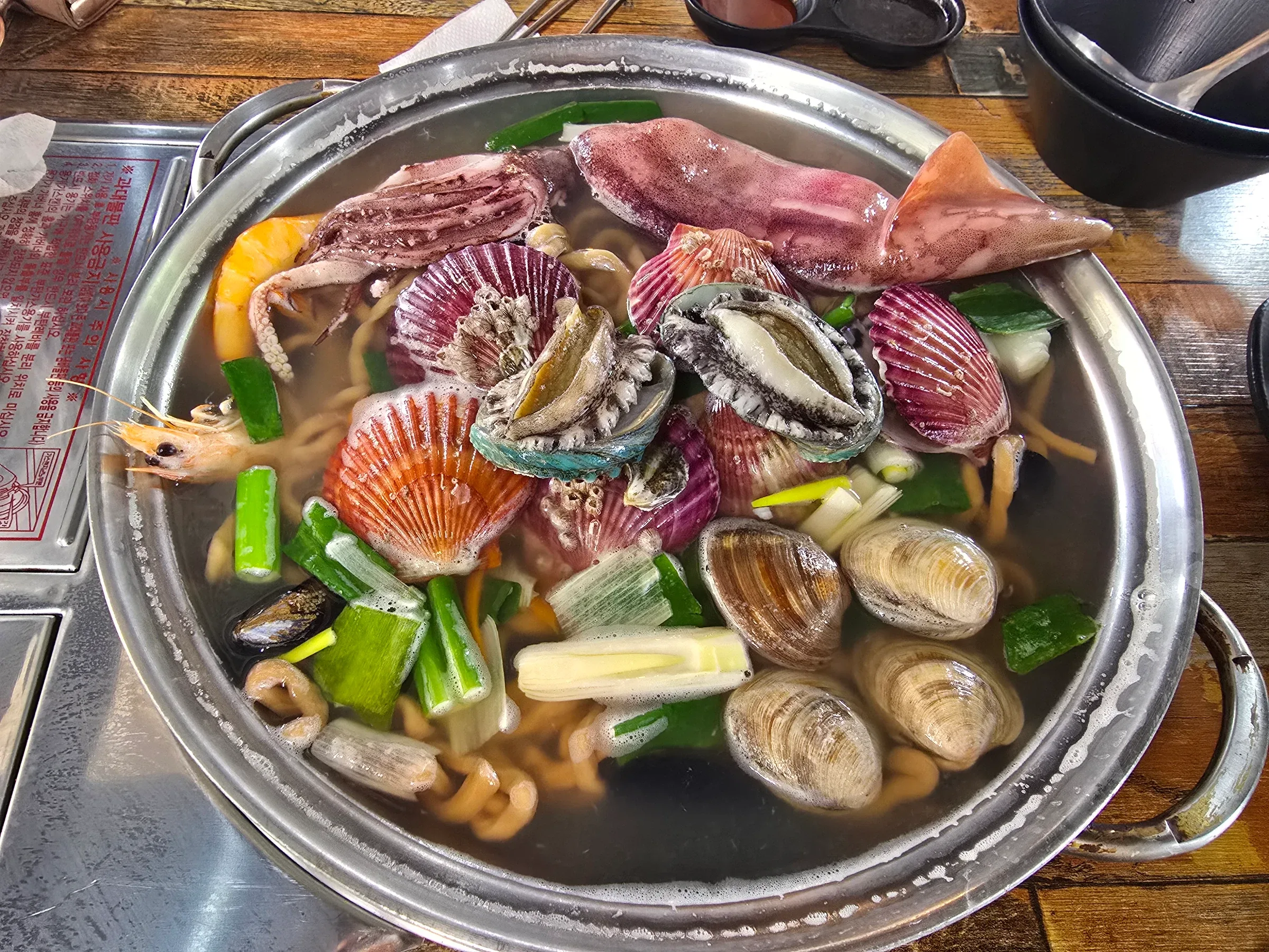 A metal pot of broth holds squid, scallops, abalone, clams, shrimp, vegetables, and noodles.