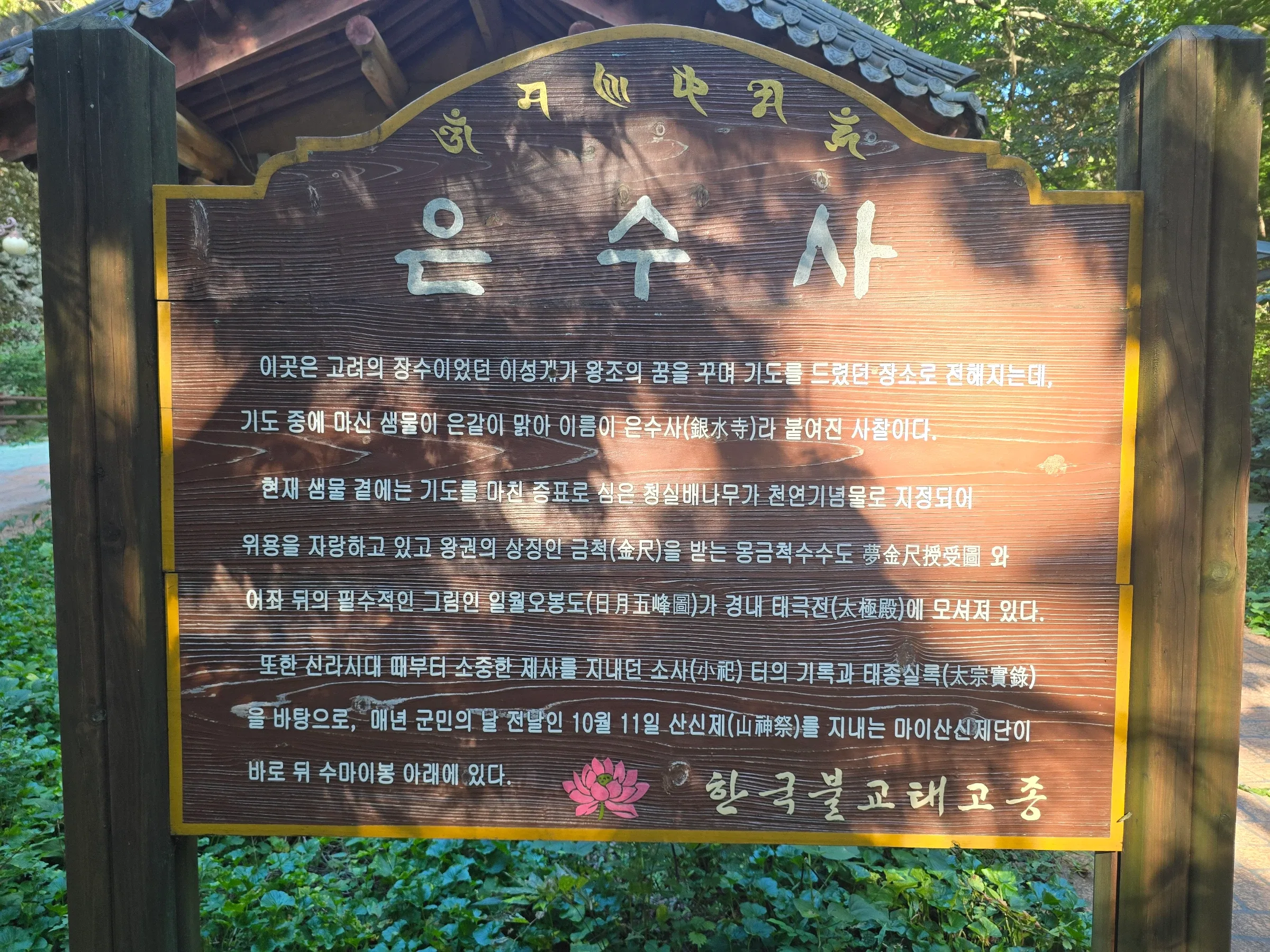 Wooden signboard at Eunsusa temple with Korean text about its spring and a Taegeukjeon hall, framed by wooden posts and green foliage.
