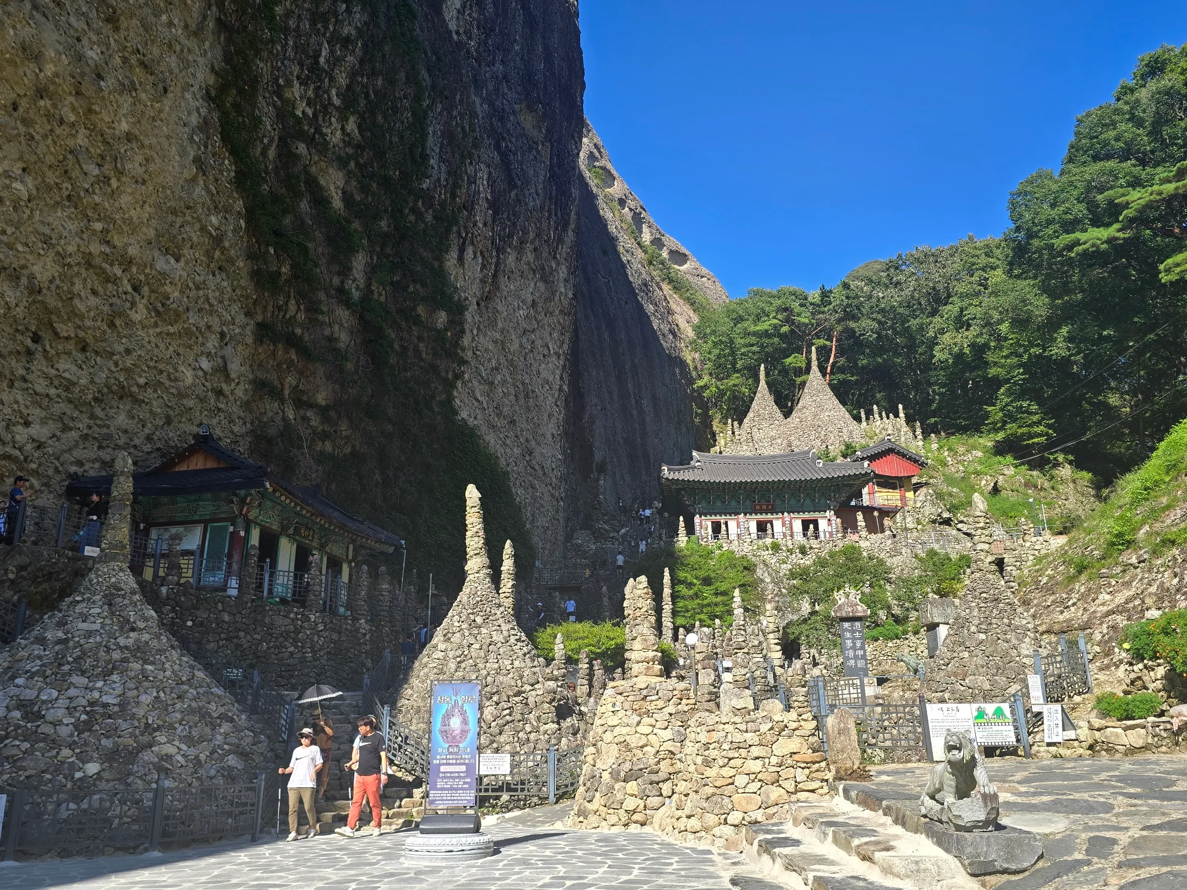 Tall rock cliff towers over a tile-roofed temple hall and cone-shaped hand-stacked stone pagodas, with visitors on stone steps and a tiger statue under a blue sky.