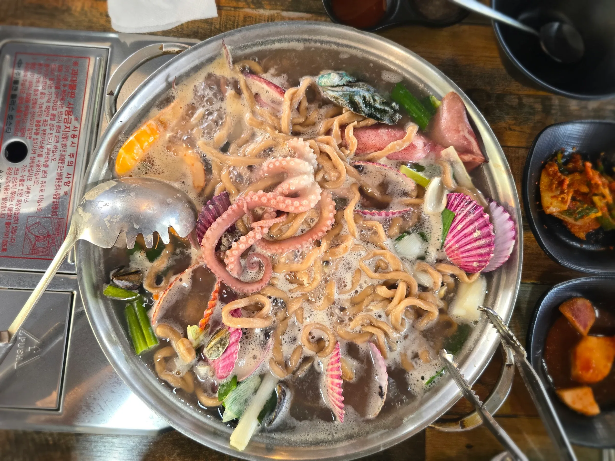 A bubbling pot of noodle soup with octopus, shellfish, scallions, and a metal ladle sits on a tabletop burner.