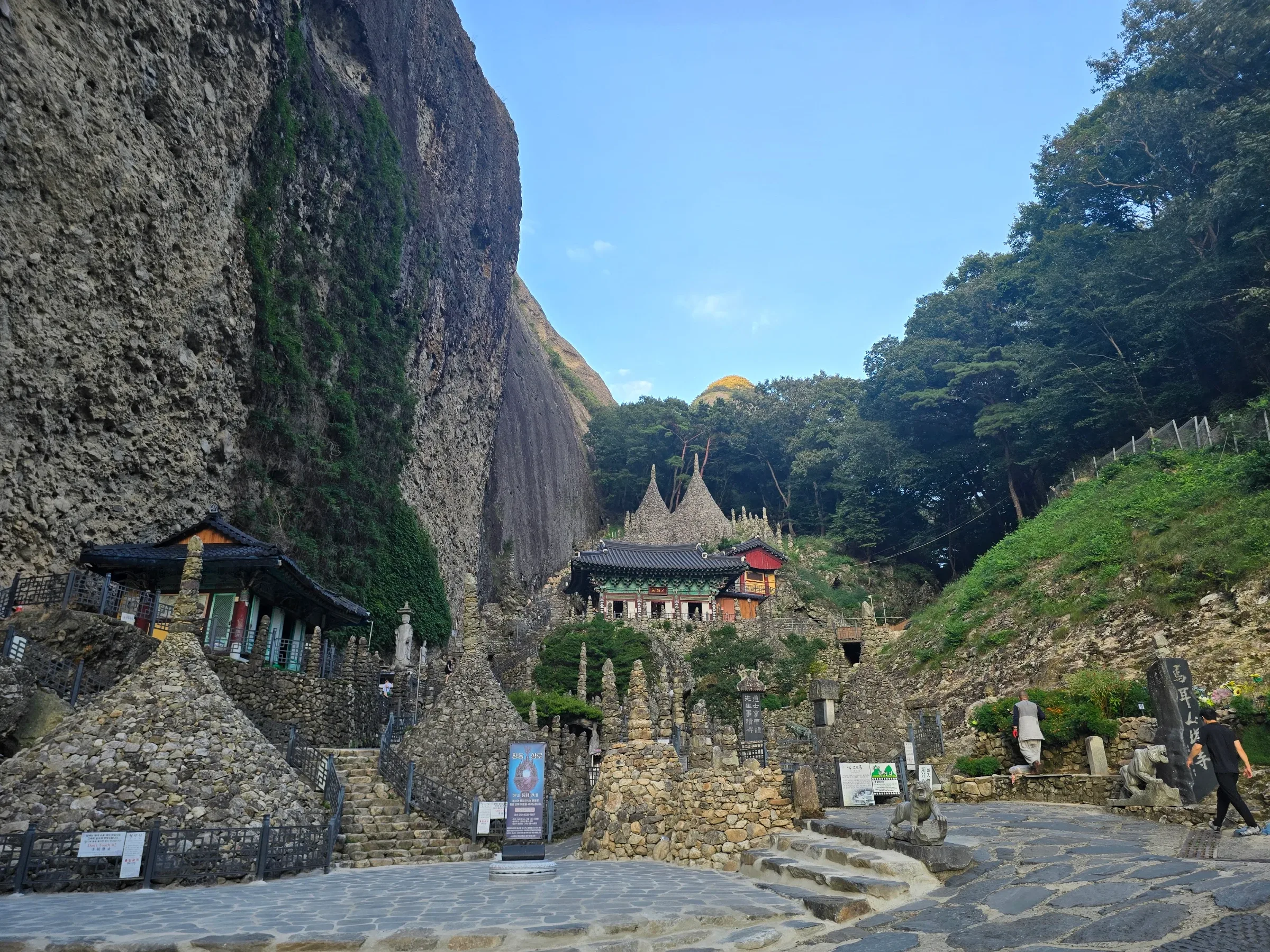 Cone-shaped hand-stacked stone towers and a tiled-roof temple hall below a huge rock cliff, with steps, stone tiger statues and two visitors.