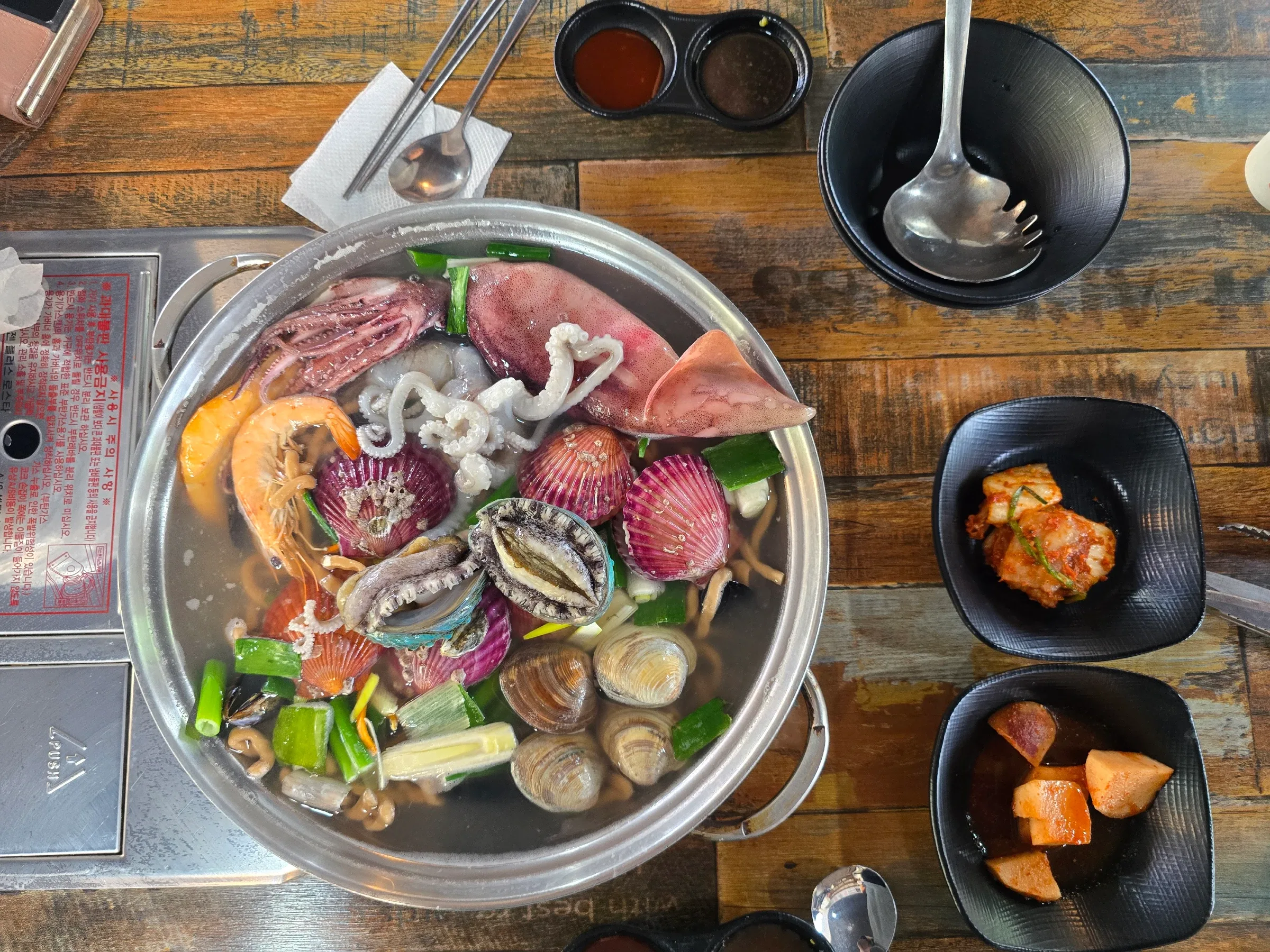 A metal pot of seafood soup with squid, shrimp, clams, scallops, and green onions sits beside small side dishes.