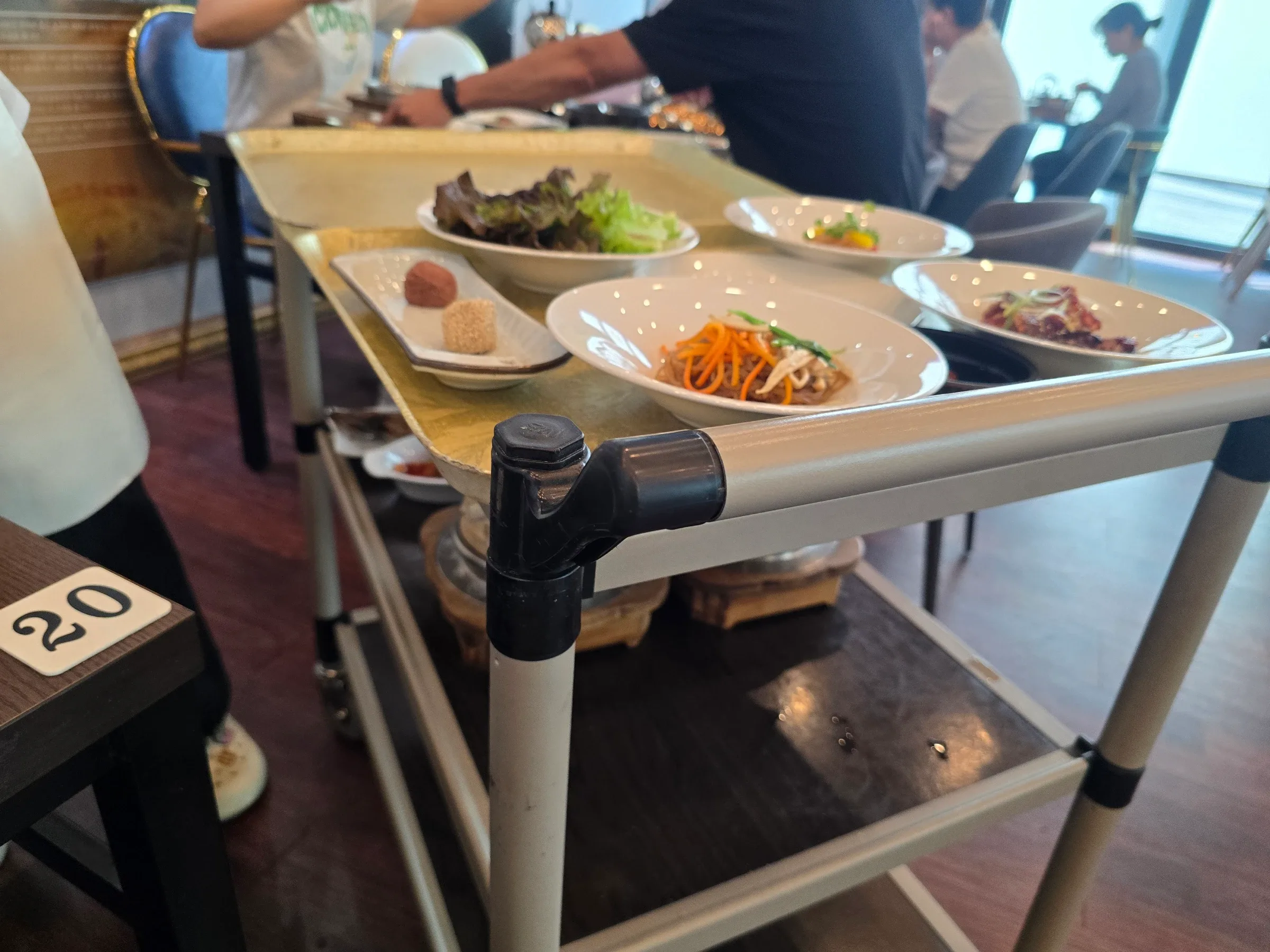Serving cart with gold trays holding white bowls of japchae, red and green lettuce, a red-sauced dish and two sweet rice balls on a plate, beside table 20.