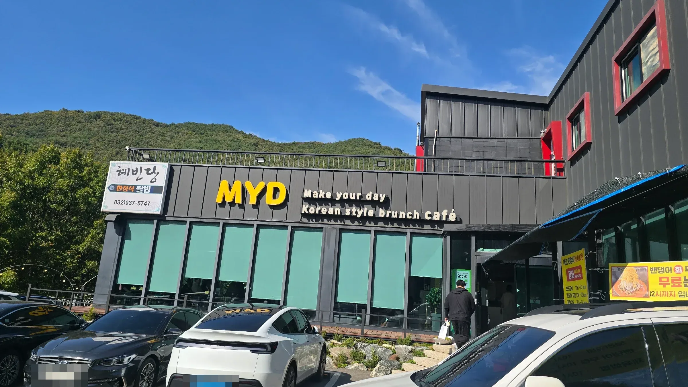 Dark-paneled MYD Korean style brunch café with green window blinds and a Hyebindang hanjeongsik sign, cars parked in front, green hill behind.
