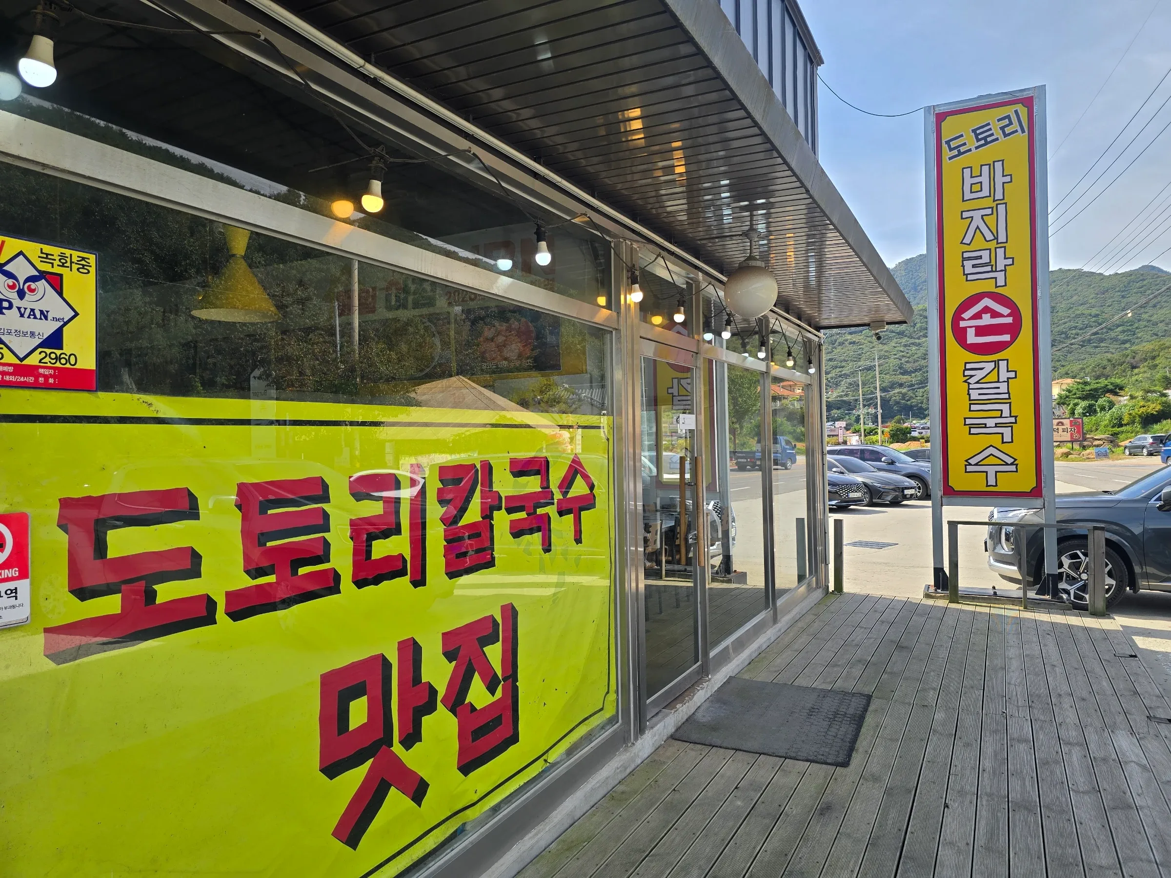 A glass-fronted restaurant with large yellow Korean signs stands beside a road, with parked cars and green hills behind it.