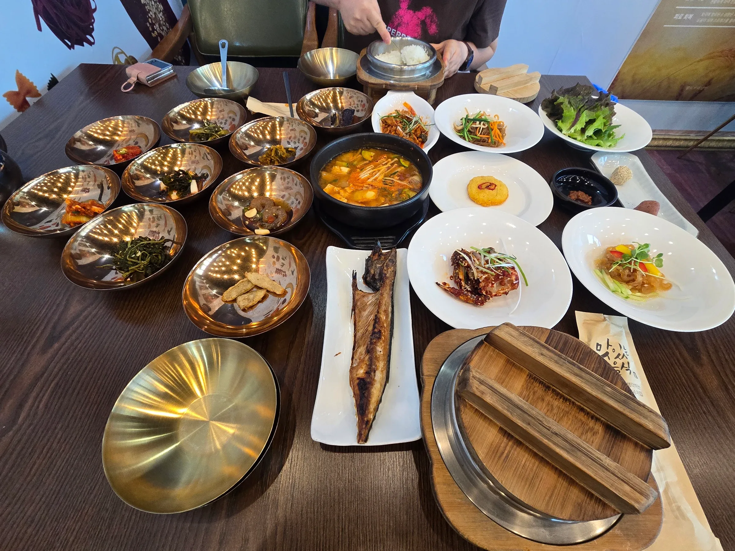 Brass bowls of banchan, a bubbling tofu stew, grilled fish, crab, jellyfish and lettuce on a dark table, with a pot of rice nearby.