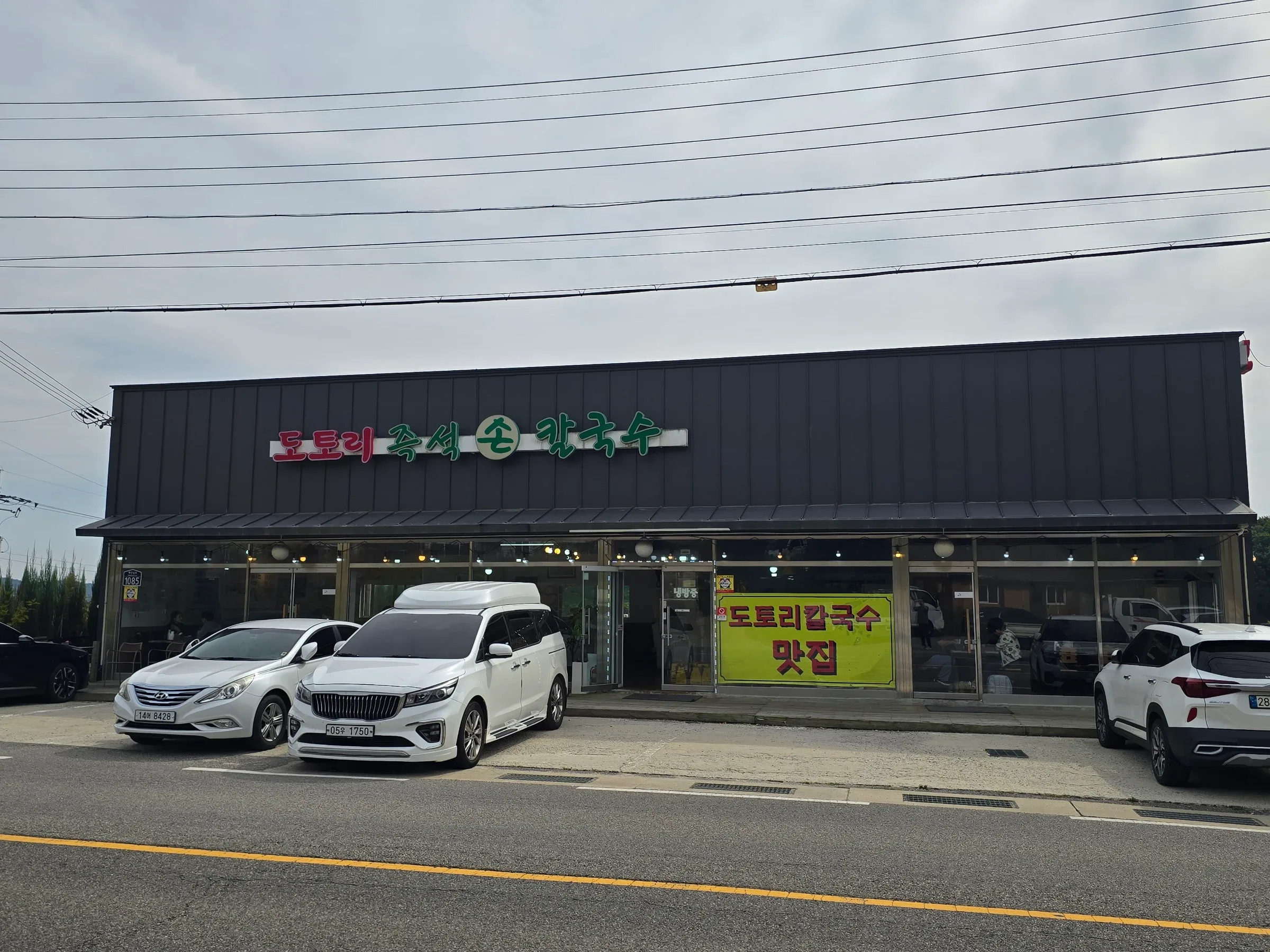 A dark-fronted restaurant with Korean signs advertising acorn kalguksu stands behind several parked cars.