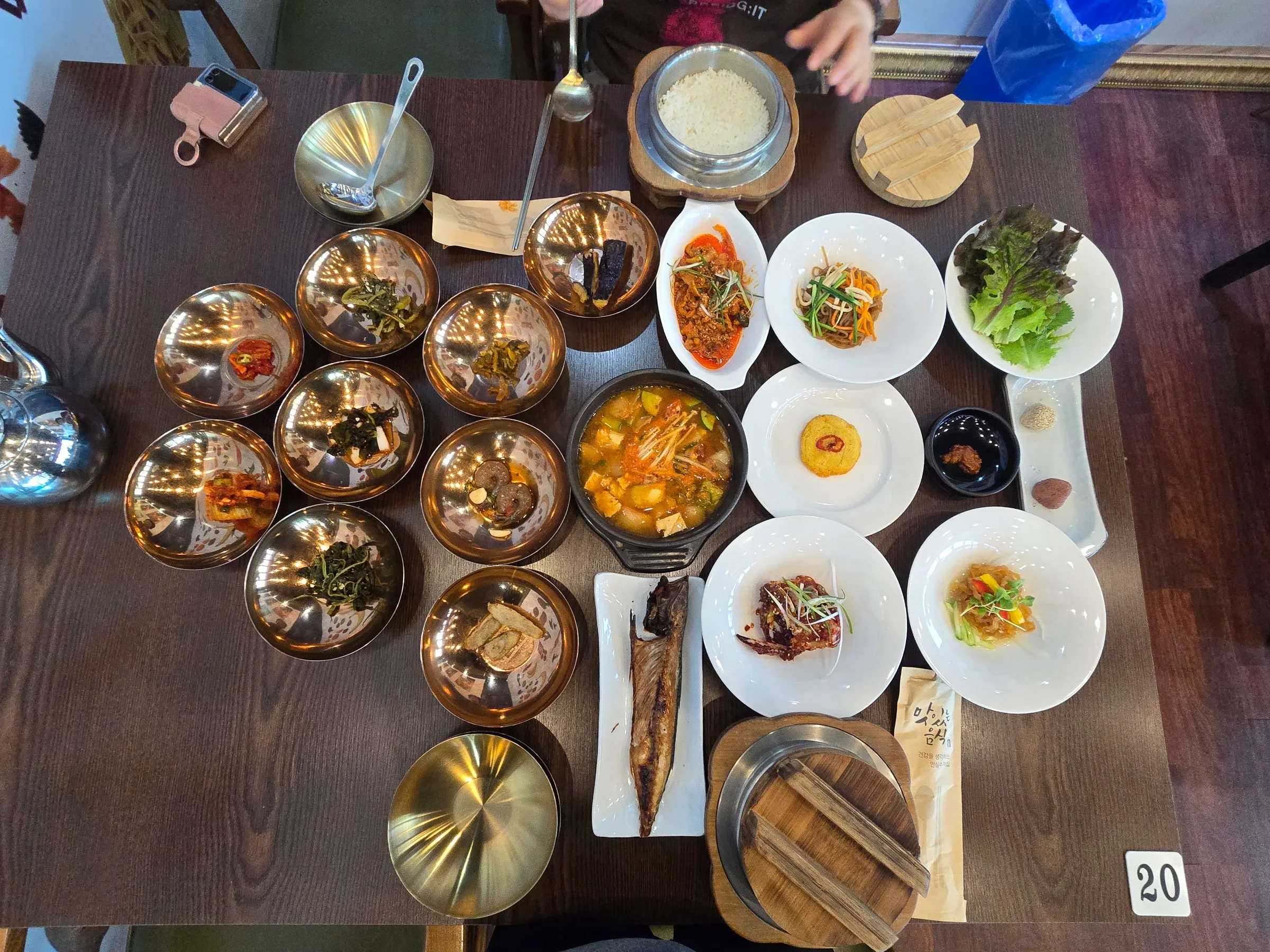 Overhead view of a dark table filled with a Korean hanjeongsik spread: brass banchan bowls, a central stew pot, white plates, grilled fish and pot rice.